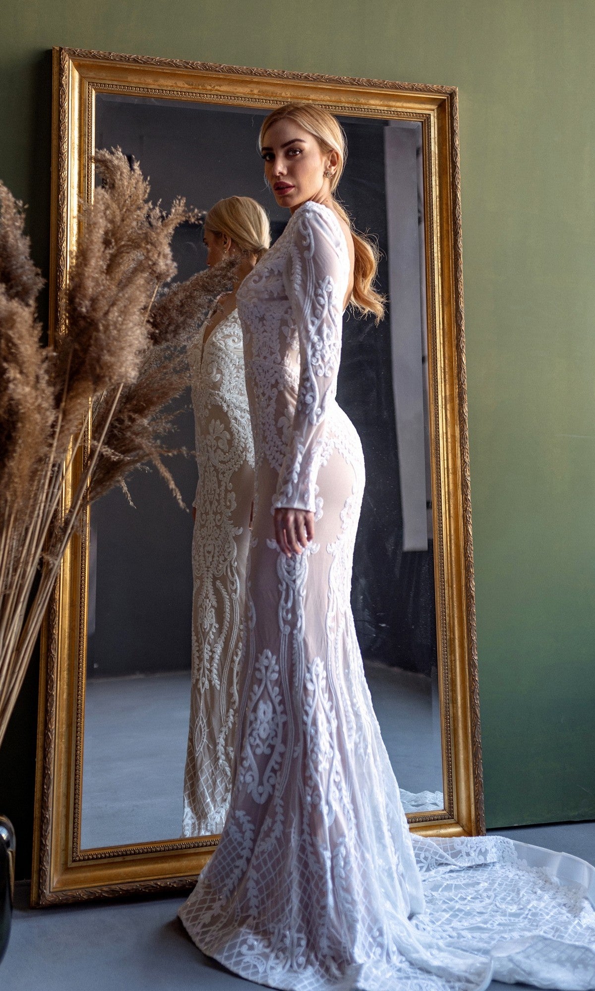 A woman wearing the Long Sleeve White Bridal Gown: Tina Holly BB021 stands before a large framed mirror, with tall dried grasses in a vase nearby.
